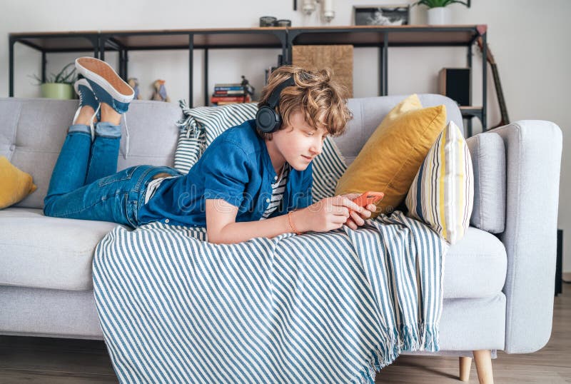 Preteen boy lying at home on cozy sofa dressed casual jeans and shirt. listening to music and chatting using wireless headphones
