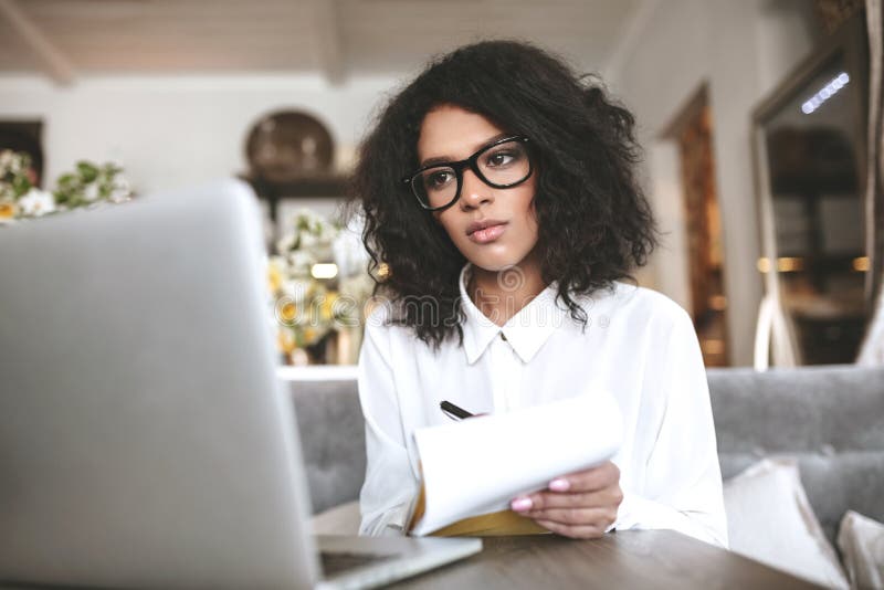 Pretty African American girl in glasses working on laptop in restaurant. Young lady with dark curly hair sitting at cafe stock photo Pretty African American girl in glasses working on laptop in restaurant. Young lady with dark curly hair sitting at cafe stock photo