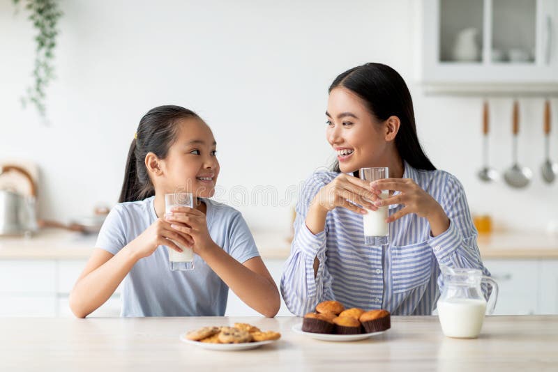 Pretty asian teen girl drinking milk with her mom, sitting at kitchen table, having pleasure conversation