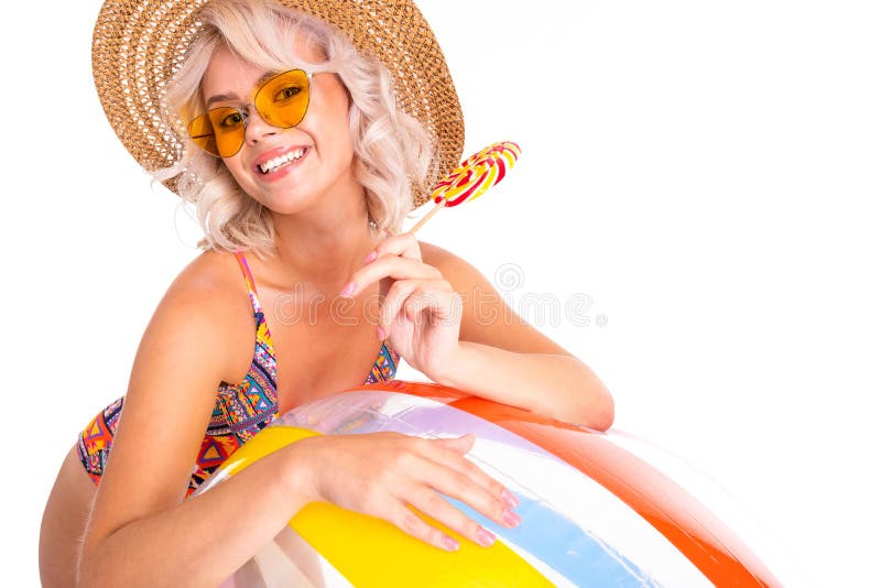 Pretty blonde caucasian female stands in swimsuit with big rubber beach colourful ball, lolipop and smiles isolated on