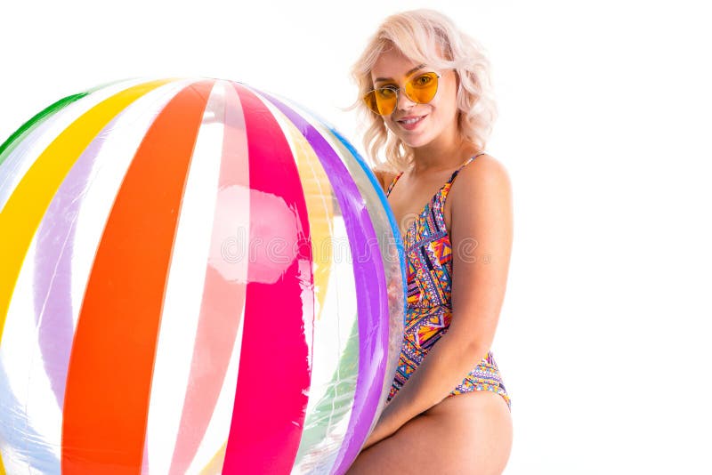 Pretty blonde caucasian female stands in swimsuit with big rubber beach colourful ball and smiles isolated on white
