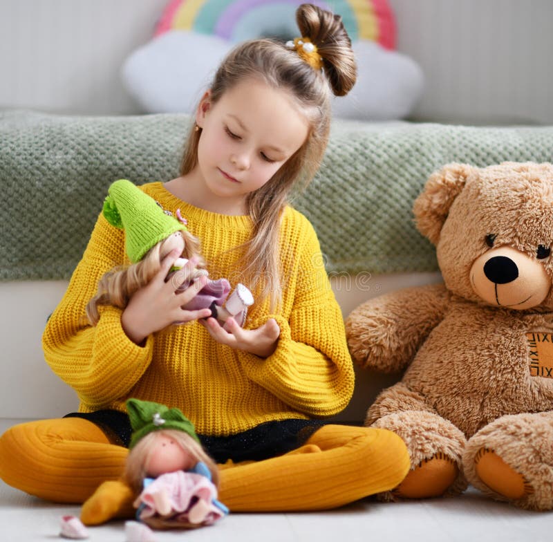 Pretty blonde small girl child in warm yellow knitted comfortable home sweater and tights sitting on floor and playing with doll