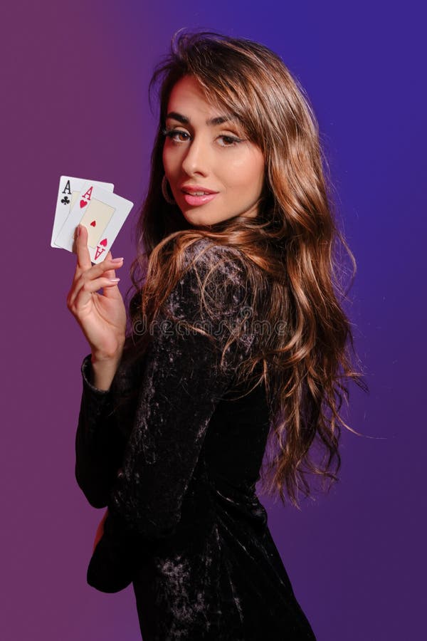 Brunette girl in black velvet dress showing two playing cards, posing sideways on coloful background. Gambling