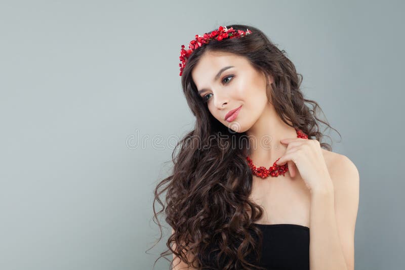 Pretty brunette woman with makeup, long curly hair and red coral necklace on gray background