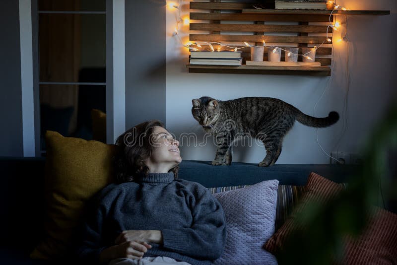 Pretty cheerful young woman 30s  years spending  time sitting with a gray cat on the sofa at home