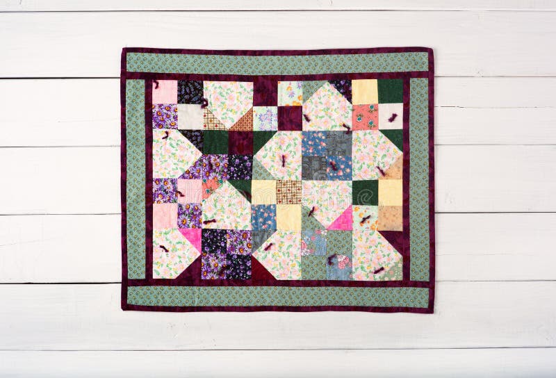 A small quilt made by me that is bordered with dark outlined green fabric with a variety of yellows, purples, blues pinks, and white colors in center area. The quilt in in center of white board background. A small quilt made by me that is bordered with dark outlined green fabric with a variety of yellows, purples, blues pinks, and white colors in center area. The quilt in in center of white board background.