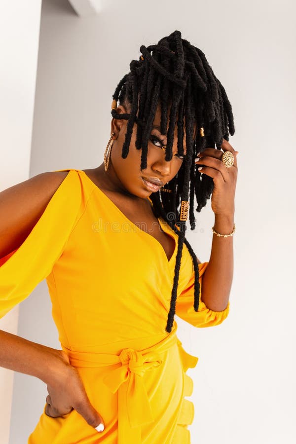 Pretty elegant woman in beautiful fashionable summer yellow dress straightens dreadlocks on white background in studio