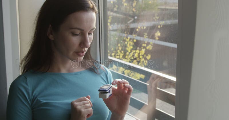 Pretty Female is Using Pulse Oximeter Checking Her Oxygen Level Standing by the Window