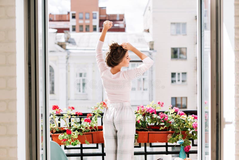 Pretty girl from back in pajamas on balcony in sunny morning. She raises hand above, looks enjoyed with closed eyes.