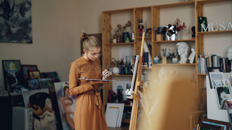 Pretty girl painter is working in workroom alone holding palette and brush and looking at canvas. Occupation, creative