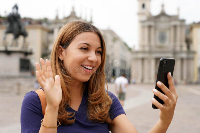 Pretty girl video calling and greets with mobile phone during her travel in Europe. Young traveler woman on landmark city square