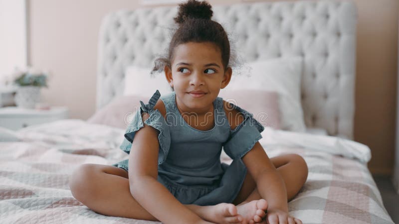 Pretty happy smiling afro american girl child with curly hair sits on bed in cozy bedroom wear blue chintz dress