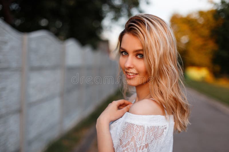 Pretty happy young beautiful blond woman in an elegant white lace blouse posing outdoors on a sunny spring day. Cute girl model