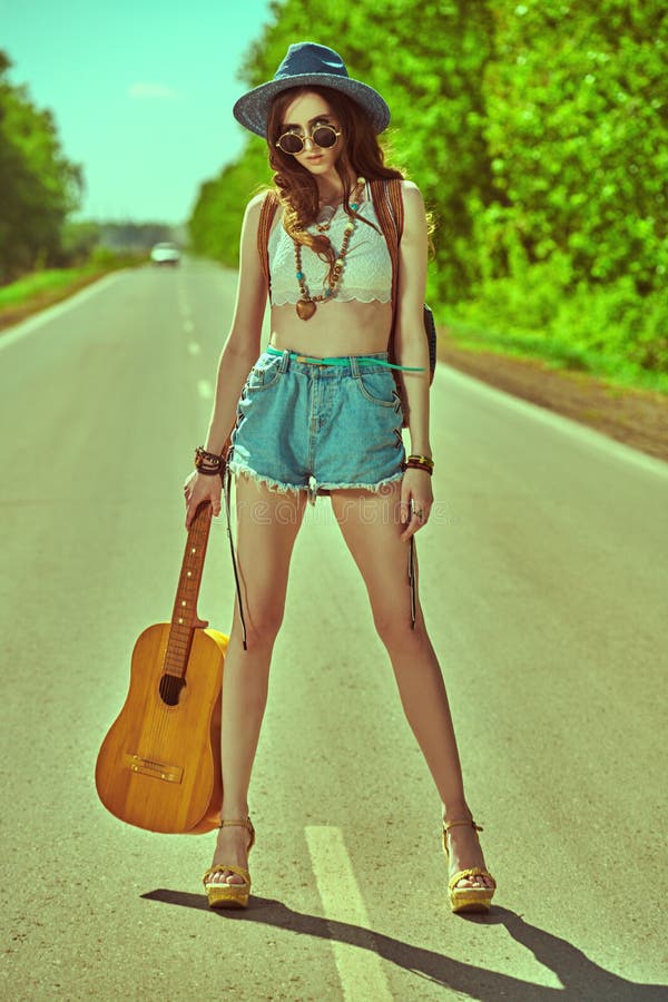 Pretty hippie girl with a guitar and a backpack hitchhiking on a highway. Spirit of freedom. Summer vacation adventure. Pretty hippie girl with a guitar and a backpack hitchhiking on a highway. Spirit of freedom. Summer vacation adventure