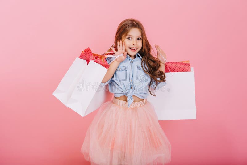 Pretty joyful young girl in tulle skirt, with long brunette hair walking with white packages on pink background. Lovely
