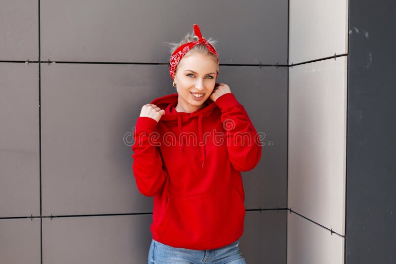 Pretty joyful young woman with a beautiful smile in a red sweater with a vintage bandana is standing near a gray wall