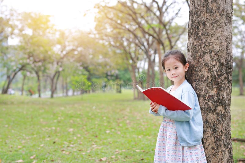 Pretty little child girl reading book in park outdoor standing lean against tree trunk in summer garden stock photography Pretty little child girl reading book in park outdoor standing lean against tree trunk in summer garden stock photography