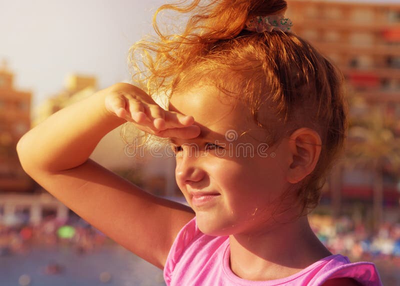 A pretty little girl looks to far away from right to left side, smiling and squinting in sunshine on city beach background. Sunset