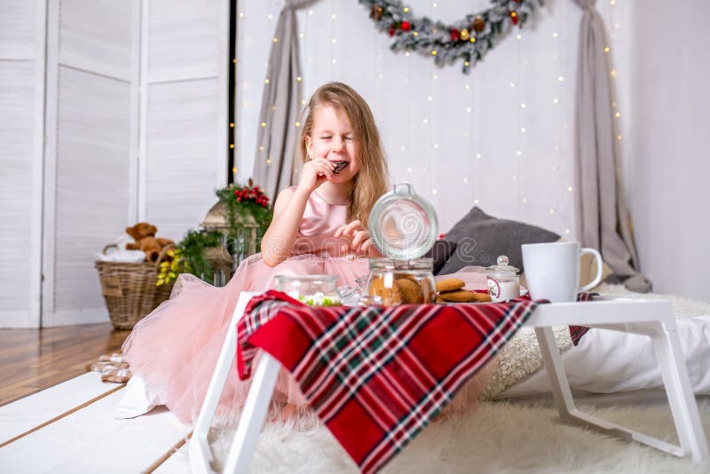 Pretty little girl 4 years old in a pink dress. Child in the Christmas room with a bed, eating candy, chocolate, cookies and drink