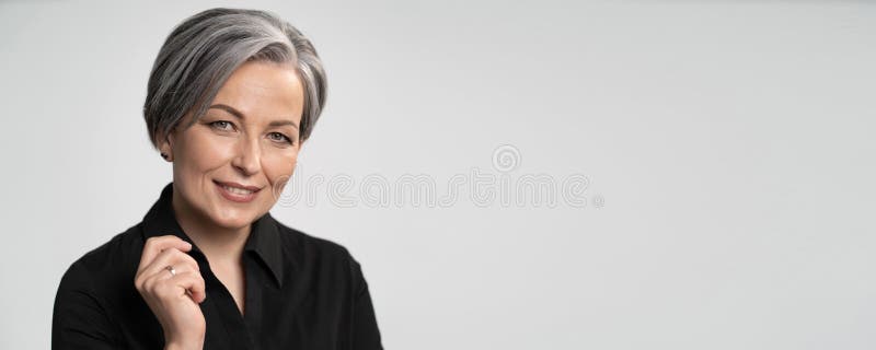 Pretty mature woman smiles with hand near face. Charming gray-haired lady in black blouse on white background. Copy