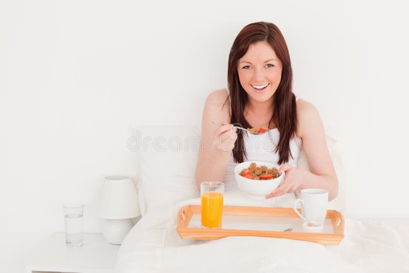 Pretty red-haired female having her breakfast while sitting on her bed