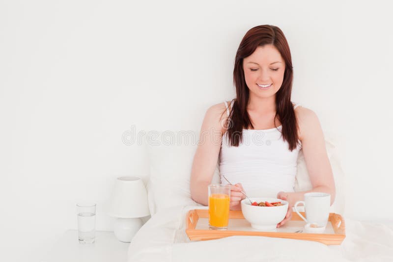 Pretty red-haired woman having her breakfast while sitting on her bed