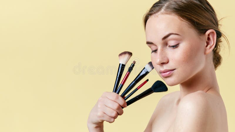 Pretty redhead teenage girl with freckles, looking down, holding diverse make up brushes. Model with light make-up.