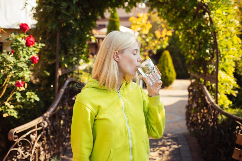 Pretty student female with blonde hair, dressed casually, having a glass of water, being happy. Outside shot of good