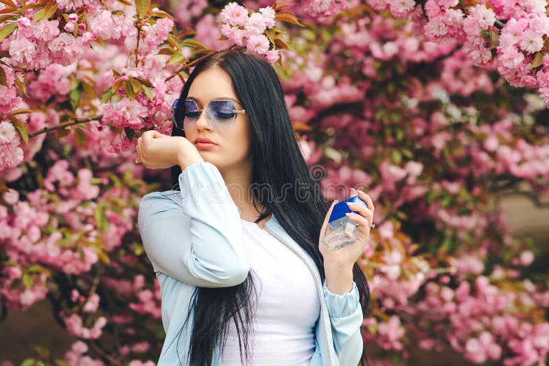 Pretty stylish girl in glasses. Pink sakura flower bloom in spring. Perfume, cosmetics concept. Beautiful woman applying perfume