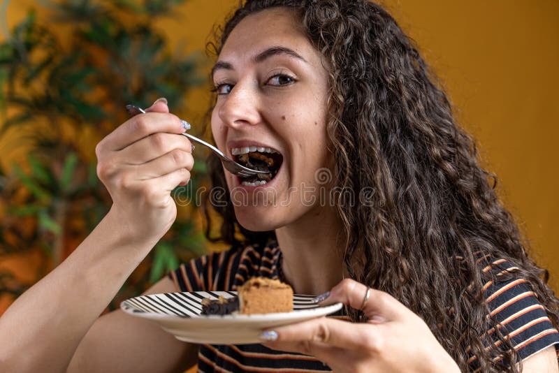 Pretty woman with curly hair sitting on a table eating a slice of chocolate cake
