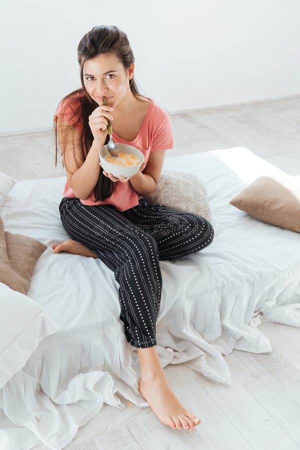 Pretty cute young woman in pajamas sitting and eating cereals with milk on bed. Pretty cute young woman in pajamas sitting and eating cereals with milk on bed