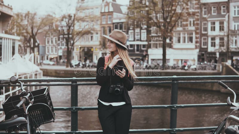 Pretty woman using smartphone on a bridge. 4K. Young lady in stylish hat with long hair and camera types a message.