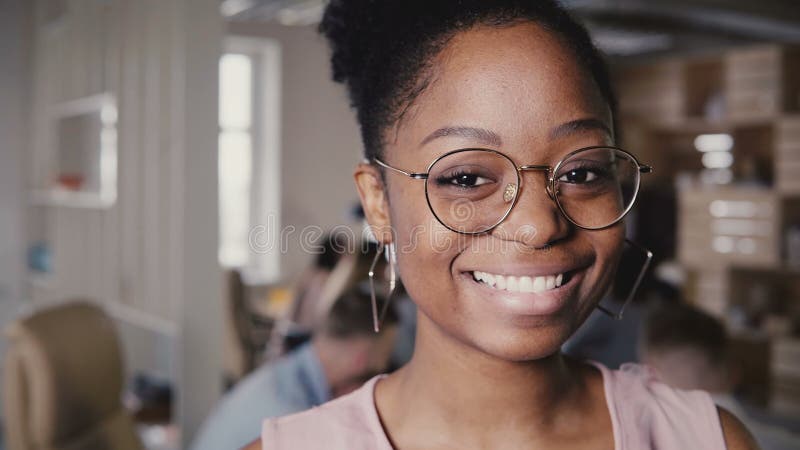 Pretty young African American happy female leader in glasses smiling at camera in modern office co-working background 4K