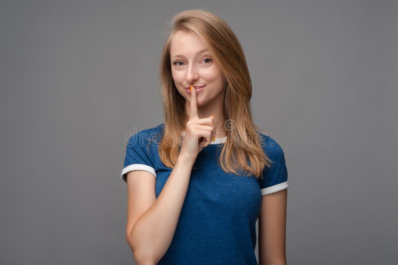 Pretty young blonde woman keeps finger on lips, making hush gesture on gray background. Shh, silence concept