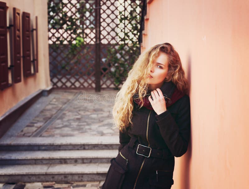 Pretty young fashion woman with long curly hair looking in camera and posing outdoor near the wall in the street background stock photography Pretty young fashion woman with long curly hair looking in camera and posing outdoor near the wall in the street background stock photography