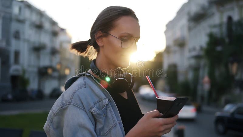 Pretty young girl in blue shirt and headphones on her neck standing on the city street and scrolling her mobile phone