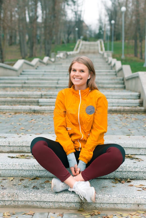 Pretty young lady, sitting on the stairs in a park si smiling at the camera and listening music in her earpods.