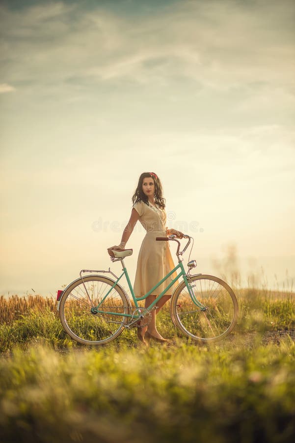 Pekná mladá smilling žena s retro bicykli v západe slnka na ceste, vintage staré časy, dievča v retro štýle na lúke