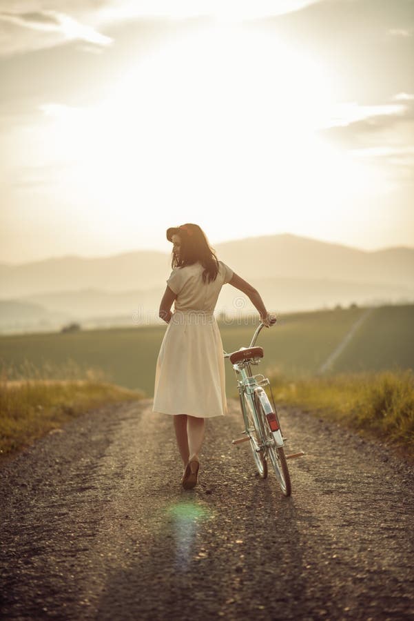 Pretty young smilling woman with retro bicycle in sunset on the road, vintage old times, girl in retro style on meadow