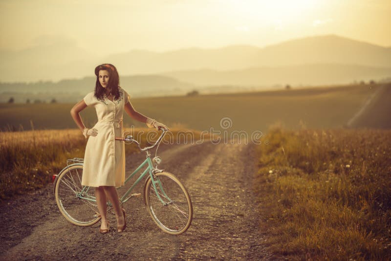 Pekná mladá smilling žena s retro bicykli v západe slnka na ceste, vintage staré časy, dievča v retro štýle na lúke