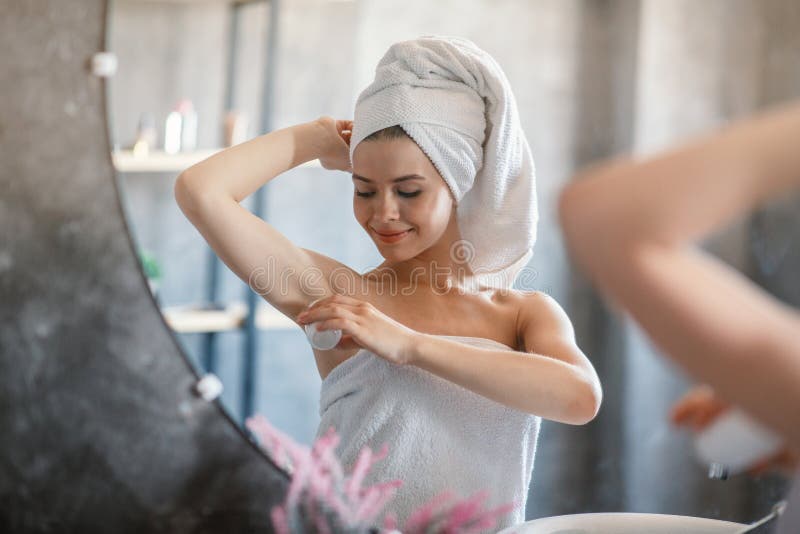 Beautiful young woman applying deodorant on armpit in front of mirror at bathroom