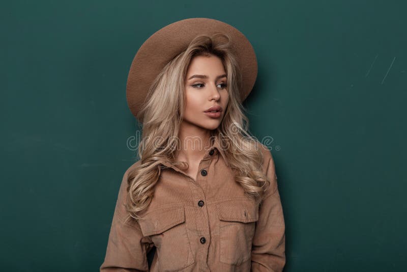 Pretty young woman in an elegant beige hat in a stylish shirt with blond curly blond hair stands in the studio