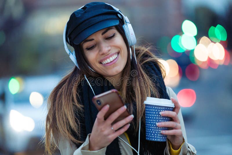 Pretty young woman listening to music with mobile phone while drinking coffee in the street at night