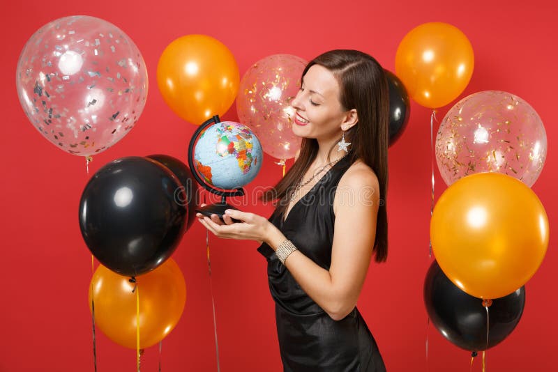Pretty young woman in little black dress celebrating, looking on world globe in hands on red background air balloons