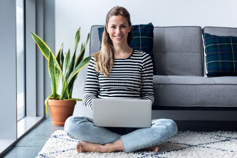 Pretty young woman looking at camera while using her laptop at home