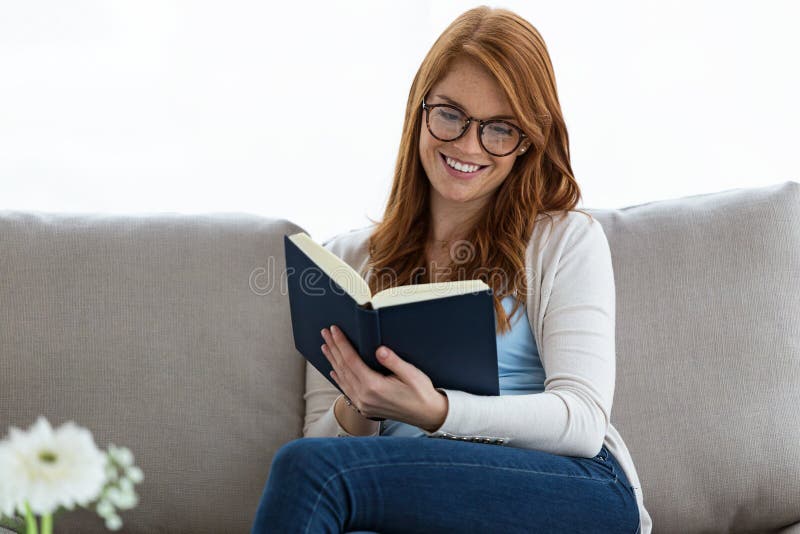 Pretty young woman reading a book while sitting on the sofa at home.