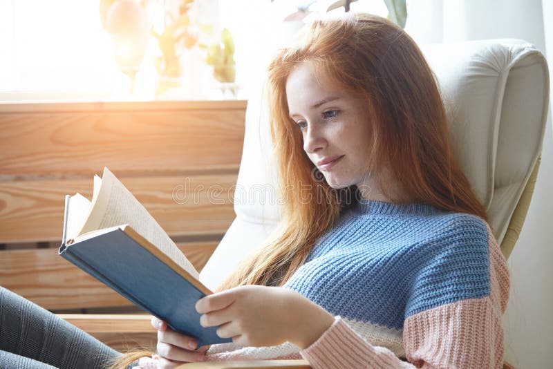 Pretty young woman with red hair sitting in comfy chair reading a book