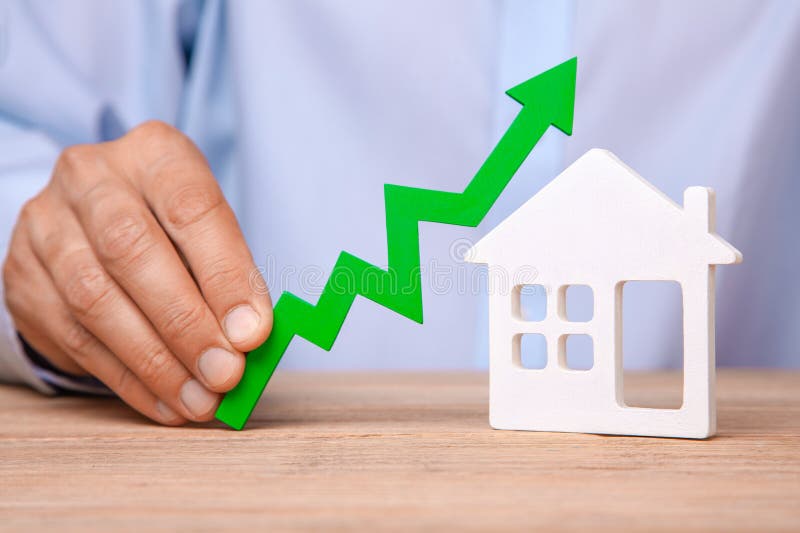 Rising house prices. Man is holding green arrow up in his hand and house on the table. Rising house prices. Man is holding green arrow up in his hand and house on the table.