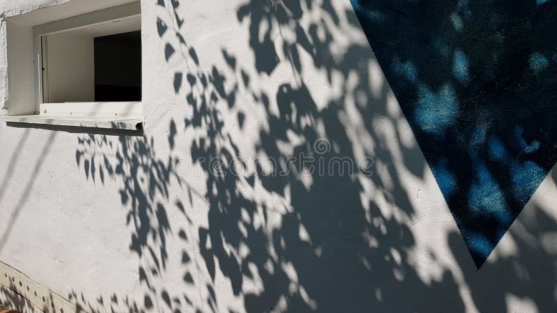 Spotted shadows from tree foliage on white stucco wall background with blue triangle painted element. Shadow patterns on house wall with window. Summer travel backdrop. Abstract background. Spotted shadows from tree foliage on white stucco wall background with blue triangle painted element. Shadow patterns on house wall with window. Summer travel backdrop. Abstract background.