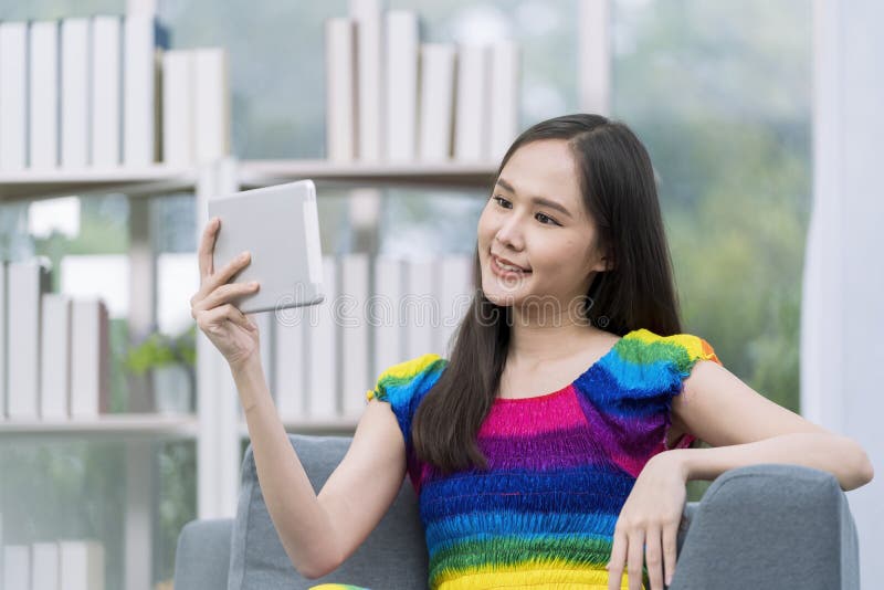 Pride Month. Young Lesbian in a rainbow dress is using a tablet on the sofa in the living room. Young woman selfie with tablet stock photos